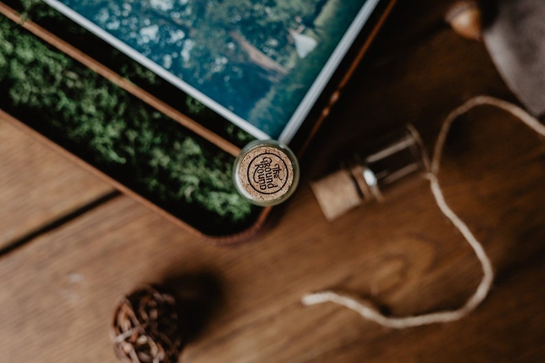 May include: A small glass bottle with a cork top. The cork has a label that reads "The Ground Round". The bottle is sitting on a wooden surface with green moss in the background.