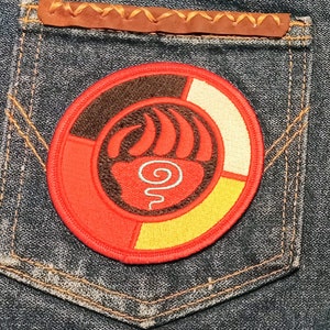 May include: A red, yellow, white, and brown embroidered patch with a stylized bear paw print and a spiral design in the center. The patch is sewn onto a blue denim pocket.