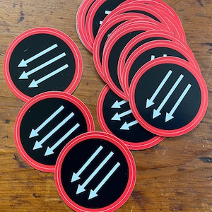 May include: A stack of black and white stickers with a red border. The stickers feature a design of three white arrows pointing to the right on a black background.