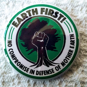Puede incluir: Un botón verde y blanco con el texto "Earth First! No Compromise in Defense of Mother Earth" y una silueta negra de un árbol con un puño en alto.