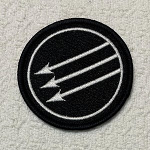 May include: A round, black patch with a white border. Three white arrows point diagonally across the center of the patch. The arrows are inside a white circle.