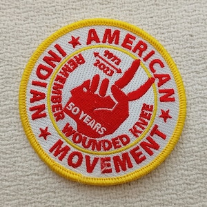 Peut inclure: Un écusson brodé blanc avec une bordure jaune. L'écusson représente une main rouge avec deux doigts levés en signe de paix. Le texte "American Indian Movement" est écrit en cercle autour de la main. Le texte "50 Years Wounded Knee" est écrit sous la main. Le texte "1973-2023" est écrit au-dessus de la main.