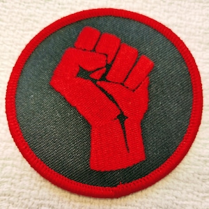 May include: Circular patch featuring a raised red fist on a dark gray background, encircled by a red border. The fist is a symbol of solidarity and resistance, commonly used in activism and social movements.