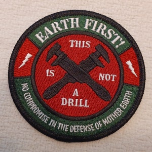 May include: A green and red embroidered patch with the text "EARTH FIRST!" in white. The patch features a pair of crossed wrenches with the text "THIS IS NOT A DRILL" and "NO COMPROMISE IN THE DEFENSE OF MOTHER EARTH".