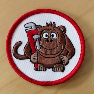 Könnte beinhalten: Ein brauner Affe mit einem roten Schraubenschlüssel, der auf einem weißen Patch mit rotem Rand gestickt ist.