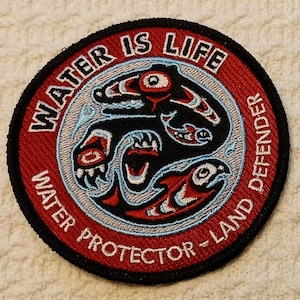 May include: A red, white, and blue embroidered patch with a stylized image of a bear and fish. The text "WATER IS LIFE WATER PROTECTOR - LAND DEFENDER" is written around the image.