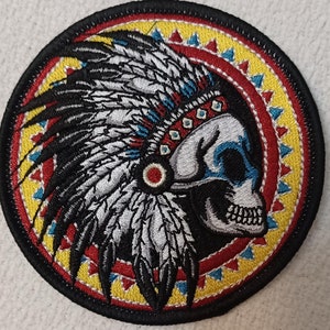 May include: Embroidered patch featuring a skull wearing a colorful Native American headdress. The skull is white with black eyes and a red nose. The headdress is black, white, red, blue, and yellow. The patch is surrounded by a red, yellow, and blue circular design.