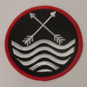Könnte beinhalten: Ein gestickter Patch in Schwarzweiß mit rotem Rand. Der Patch zeigt zwei gekreuzte Pfeile, die nach links und rechts zeigen, und darunter ein weißes Wellenmuster.