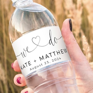 Könnte beinhalten: Klare Plastikwasserflasche mit weißem Etikett. Auf dem Etikett steht "we do" mit einem Herz, den Namen "KATE + MATTHEW" und dem Datum "August 23, 2024". Die Flasche wird vor einem Hintergrund aus hohem, trockenem Gras gehalten.