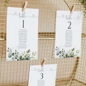 Könnte beinhalten: Drei weiße Karten mit grünen Blattwerk-Designs und dem Text "Tisch" gefolgt von einer Nummer, 1, 2 und 3. Jede Karte listet eine Reihe von Namen auf.