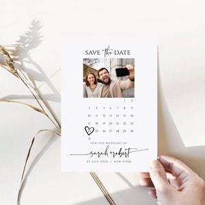 May include: Save the Date card with a calendar featuring a photo of a couple. The date 25 is circled with a heart. The text reads "Save the Date" and "For the wedding of Sarah + Robert" with the date "25 July 2024" and location "New York".
