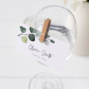 Puede incluir: Una tarjeta de lugar blanca con un diseño de eucalipto acuarela y el texto "Olivia Smith Mesa Uno" unido a una copa de vino con una pinza de madera.