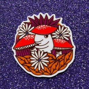 May include: A sticker featuring a cartoon illustration of three red and white mushrooms with white spots, surrounded by white flowers and purple and orange leaves.