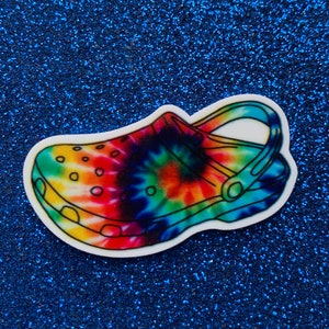 May include: A tie-dye patterned clog with a white background and a black outline. The clog has a strap across the top and a hole pattern on the sides.