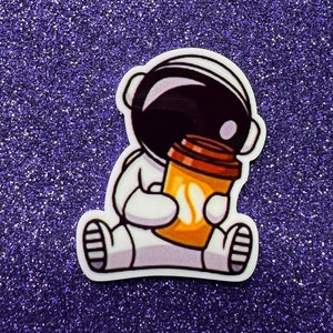 May include: A cartoon astronaut wearing a white spacesuit and a black helmet is holding a brown and orange coffee cup. The astronaut is sitting down with its legs crossed.