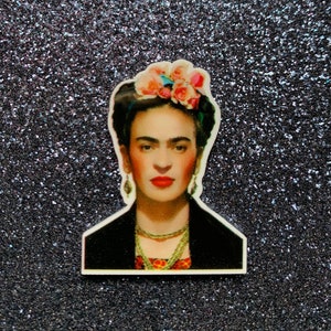 May include: A colorful portrait of Frida Kahlo with a flower crown, black jacket, and layered necklaces. The background is a sparkly silver.