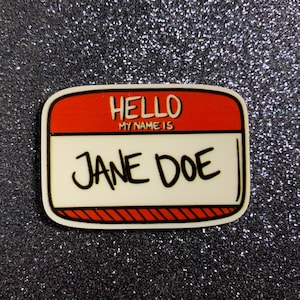 May include: A name tag with a red top section that reads "HELLO MY NAME IS" and a white bottom section with the name "JANE DOE" in black. The name tag has a black outline and is set against a glittery, dark background.