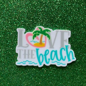 May include: A white sticker with the words "I LOVE THE beach" in gray and teal. The word "LOVE" has a pink heart with a palm tree and island graphic. The sticker is on a green glitter background.