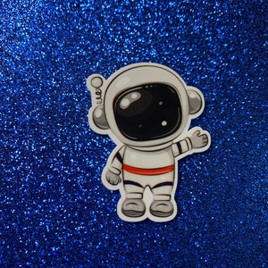 May include: A white astronaut sticker with a red stripe on its suit, against a blue glitter background.