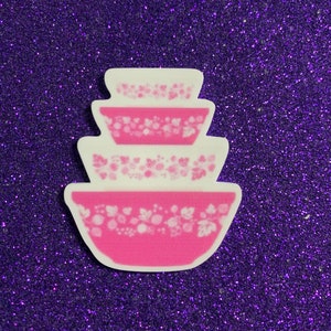 May include: A stack of three pink and white mixing bowls with a floral pattern. The bowls are arranged in a nested configuration, with the largest bowl at the bottom and the smallest at the top. The background is a sparkly purple.