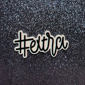 May include: A white sticker with black text that says "#extra" on a sparkly black background.