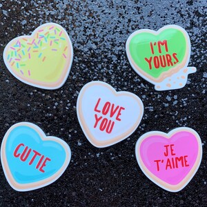 May include: Five heart-shaped cookies with different messages. The cookies are decorated with sprinkles, icing, and different colored backgrounds. The messages on the cookies are "I'm Yours", "Love You", "Cutie", "Je T'aime", and a cookie with yellow icing and sprinkles.