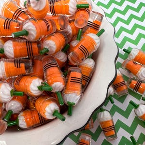 K&ouml;nnte beinhalten: Eine wei&szlig;e Sch&uuml;ssel gef&uuml;llt mit Miniatur-Bechern aus orangefarbenem und wei&szlig;em Kunststoff mit gr&uuml;nen Strohhalmen. Die Becher sind in einem zuf&auml;lligen Muster angeordnet und einige fallen aus der Sch&uuml;ssel auf eine gr&uuml;n-wei&szlig; karierte Oberfl&auml;che.
