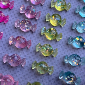 May include: Assortment of iridescent candy-shaped resin charms in pink, blue, green, and yellow colors. The charms have a glittery finish and are arranged in rows on a purple background.