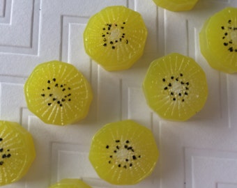 May include: A close-up of ten yellow kiwi-shaped soaps with black seeds. The soaps are arranged in a row on a white surface.