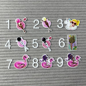 May include: A felt board displays decorative embellishments, including numbers 1-9. The embellishments feature a variety of designs, such as a lollipop, a woman on a donut, a woman on a unicorn float, and a flamingo float.
