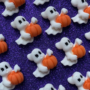 May include: A group of white ghost figurines holding orange pumpkins. The ghosts have black eyes and mouths. The pumpkins are a bright orange color. The figurines are on a purple sparkly background.