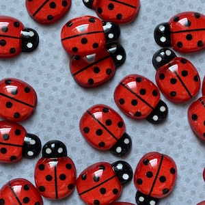 May include: A collection of red ladybug-shaped embellishments with black spots and black heads. These decorative wooden ladybugs are scattered across a light gray surface, showcasing their vibrant colors and playful design. Ideal for craft projects.