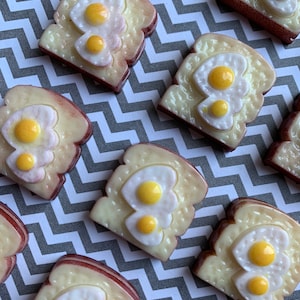 May include: A set of nine resin food charms shaped like toast with fried eggs. The charms are yellow, white, and brown. They are arranged in a staggered pattern on a gray and white chevron background.