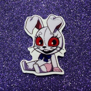 May include: A white plush toy rabbit with red eyes and a pink and white dress. The rabbit is sitting with its legs crossed and has a big smile.