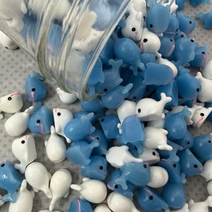 Puede incluir: Una colecci&oacute;n de peque&ntilde;as figuras en forma de ballena en tonos blanco y azul claro, derram&aacute;ndose de un recipiente de vidrio transparente. Las figuras tienen ojos negros y detalles rosas, creando una est&eacute;tica l&uacute;dica y caprichosa. Estas ballenas en miniatura est&aacute;n dispersas.