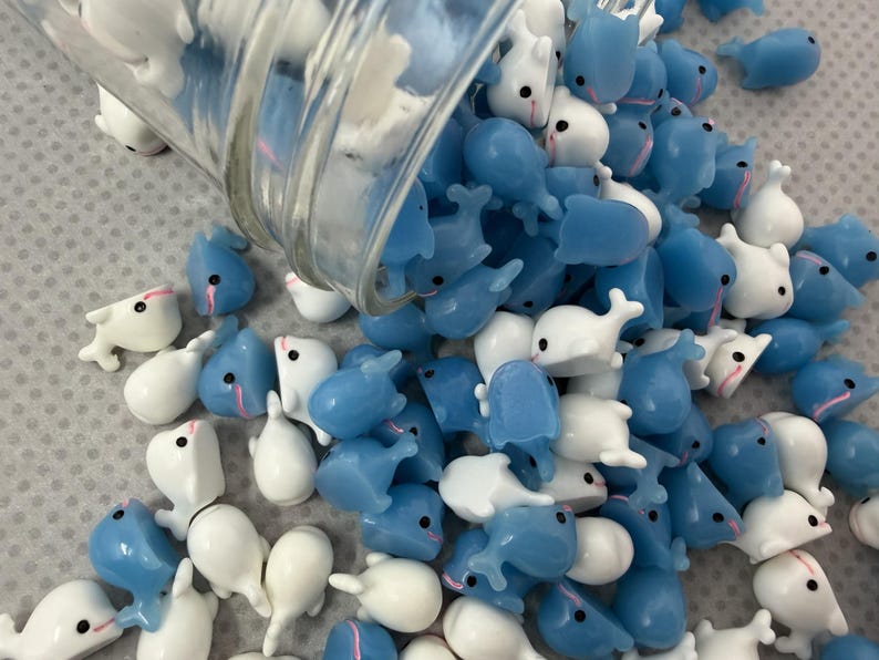 Puede incluir: Una colecci&oacute;n de peque&ntilde;as figuras en forma de ballena en tonos blanco y azul claro, derram&aacute;ndose de un recipiente de vidrio transparente. Las figuras tienen ojos negros y detalles rosas, creando una est&eacute;tica l&uacute;dica y caprichosa. Estas ballenas en miniatura est&aacute;n dispersas.