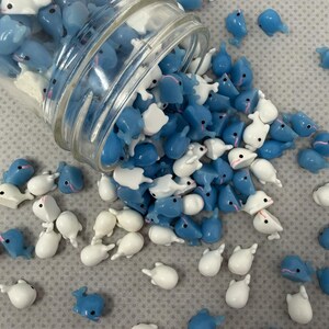 Puede incluir: Un tarro de cristal volcado, derramando una colecci&oacute;n de peque&ntilde;os amuletos con forma de ballena azules y blancos. Los amuletos est&aacute;n esparcidos sobre una superficie gris claro. Algunos son azules, otros blancos y algunos tienen acentos rosas.