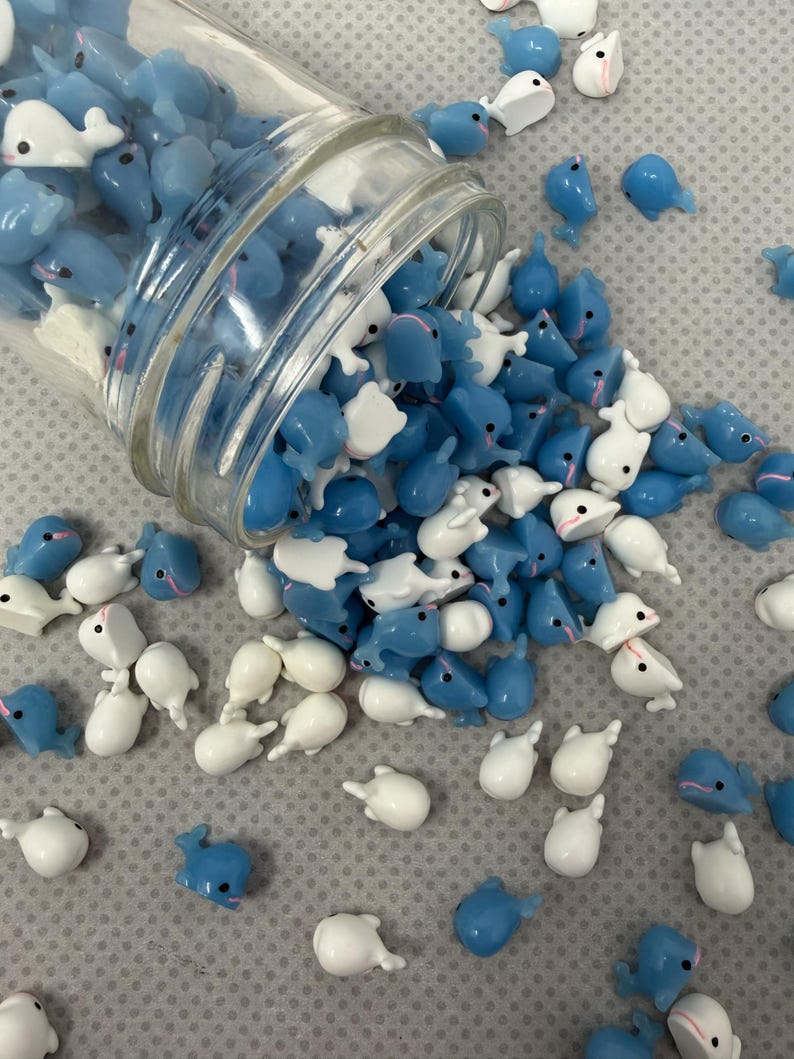 Puede incluir: Un tarro de cristal volcado, derramando una colecci&oacute;n de peque&ntilde;os amuletos con forma de ballena azules y blancos. Los amuletos est&aacute;n esparcidos sobre una superficie gris claro. Algunos son azules, otros blancos y algunos tienen acentos rosas.