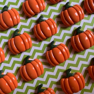 May include: A collection of 15 orange pumpkin shaped embellishments with green stems. The pumpkins are arranged in rows on a green and white chevron patterned surface.