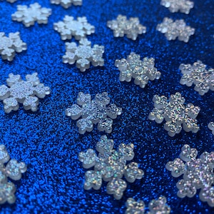 May include: A blue background covered in glitter with white snowflake-shaped ornaments scattered across the surface.