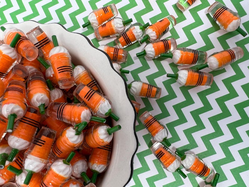 K&ouml;nnte beinhalten: Eine Sch&uuml;ssel gef&uuml;llt mit Miniatur-Karottenbechern in Orange und Wei&szlig; mit gr&uuml;nen Strohhalmen. Die Becher sind in einem zuf&auml;lligen Muster angeordnet und einige fallen aus der Sch&uuml;ssel.