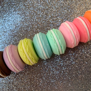 May include: A row of colorful macarons in shades of brown, purple, yellow, blue, green, pink, and orange. The macarons are arranged in a line from left to right, with the brown macaron on the left and the orange macaron on the right.