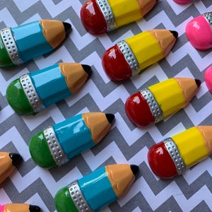 May include: Assortment of colorful pencil-shaped ornaments with a silver band and eraser. The pencils are in shades of blue, green, yellow, pink, and red.