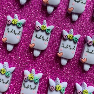 May include: A collection of light purple unicorn-shaped embellishments with closed eyes, eyelashes, and a small heart. Each unicorn has a colorful floral crown and a popsicle stick base. The background is a sparkly, bright pink surface.