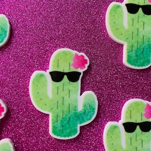 May include: Four green cactus stickers with black sunglasses and pink flowers. The cactus stickers are on a pink glitter background.