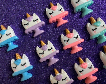 May include: Ten unicorn cake charms in a variety of colours, including pink, purple, blue, and white. Each charm features a unicorn with a gold horn and a seashell on its head. The charms are ideal for adding a whimsical touch to crafts, jewellery, and other projects.