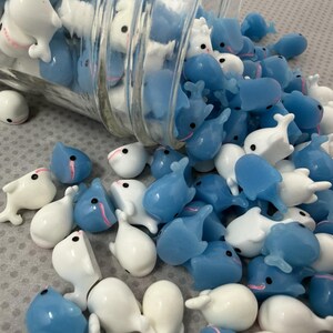 Puede incluir: Una colecci&oacute;n de peque&ntilde;as figuras con forma de ballena en blanco y azul claro, algunas derram&aacute;ndose de un frasco de vidrio transparente. Las ballenas tienen ojos negros y detalles en rosa. Las figuras est&aacute;n esparcidas sobre una superficie gris claro.