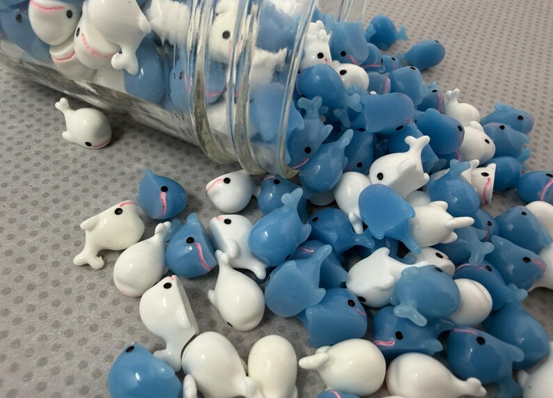 Puede incluir: Una colecci&oacute;n de peque&ntilde;as figuras con forma de ballena en blanco y azul claro, algunas derram&aacute;ndose de un frasco de vidrio transparente. Las ballenas tienen ojos negros y detalles en rosa. Las figuras est&aacute;n esparcidas sobre una superficie gris claro.