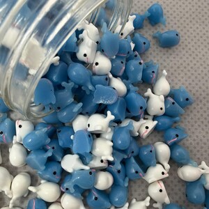 Puede incluir: Un tarro de cristal transparente volcado, derramando una multitud de peque&ntilde;as figuras con forma de ballena. Las figuras son una mezcla de azul claro y blanco, con ojos negros y detalles en rosa. El fondo es una superficie texturizada de color gris claro.