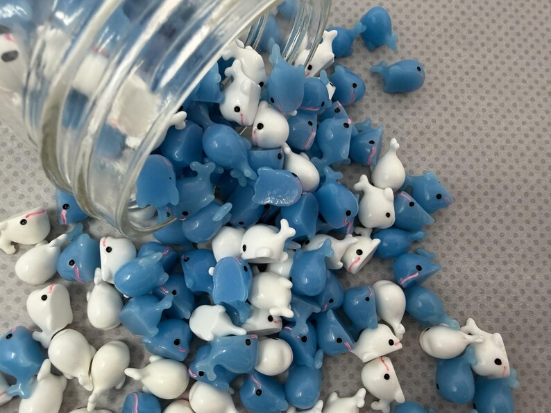 Puede incluir: Un tarro de cristal transparente volcado, derramando una multitud de peque&ntilde;as figuras con forma de ballena. Las figuras son una mezcla de azul claro y blanco, con ojos negros y detalles en rosa. El fondo es una superficie texturizada de color gris claro.